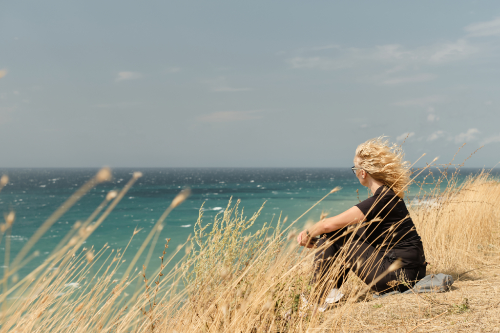 adult woman sits high bank overlooking ocean her hair blowing wind